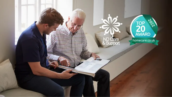 A carer and a man sit on a bench and read a book. Next to them is the logo for ND Care & Support and the logo for Homecare.co.uk's Top 20 Home Care Provider award.